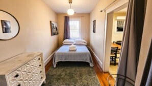 A narrow room with a bed, a dresser, and a window with curtains. - The Terrace (Charlotte)