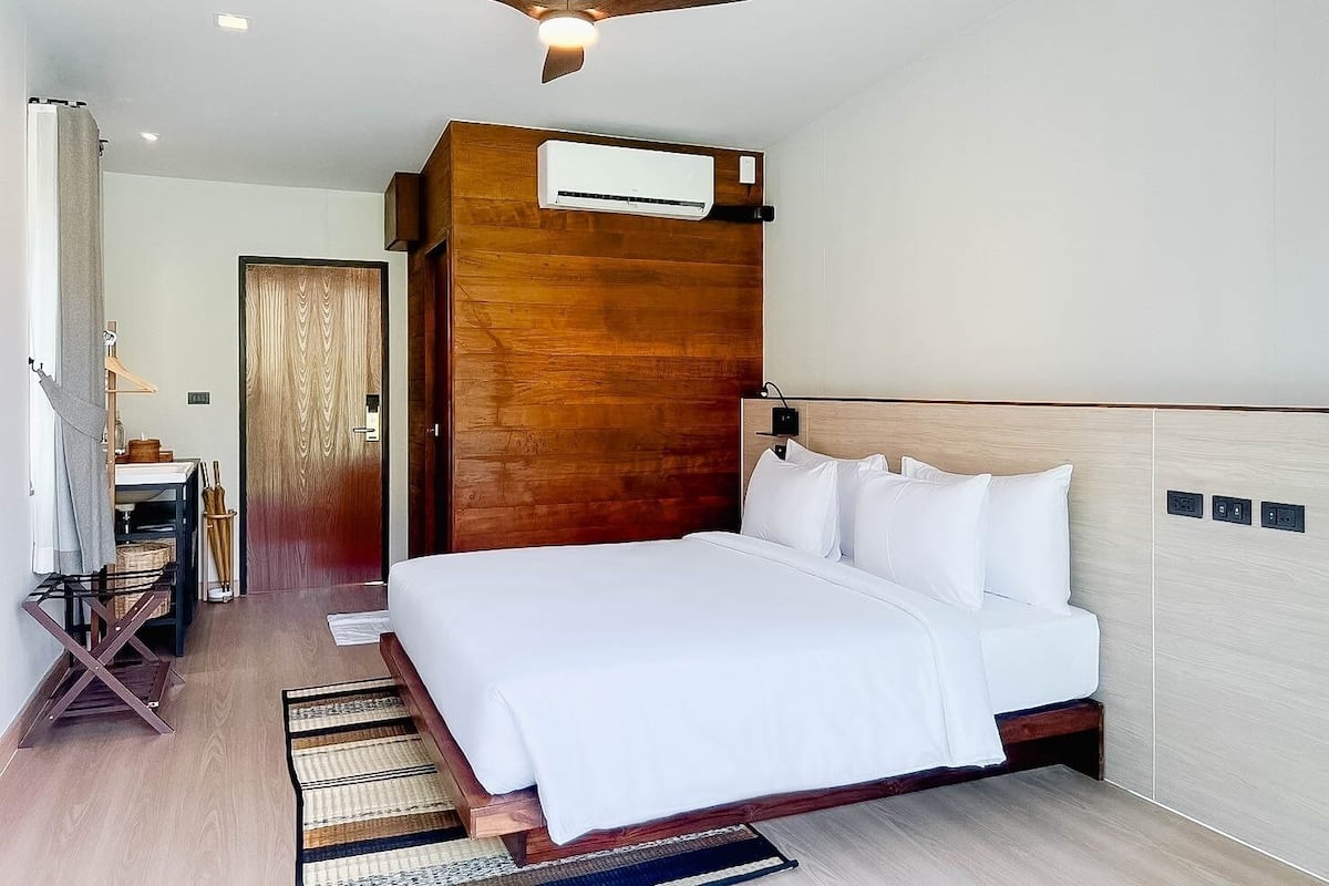 A modern hotel room with a wooden headboard, a large bed with white linens, a ceiling fan, and an air conditioning unit.