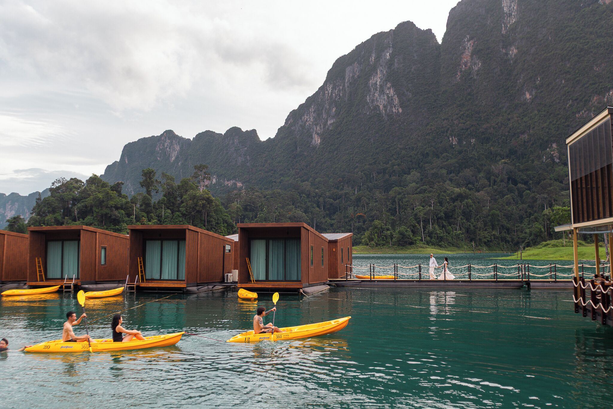 Un plan d’eau agrémenté de structures flottantes en bois, de kayaks, et dominé par un paysage montagneux.