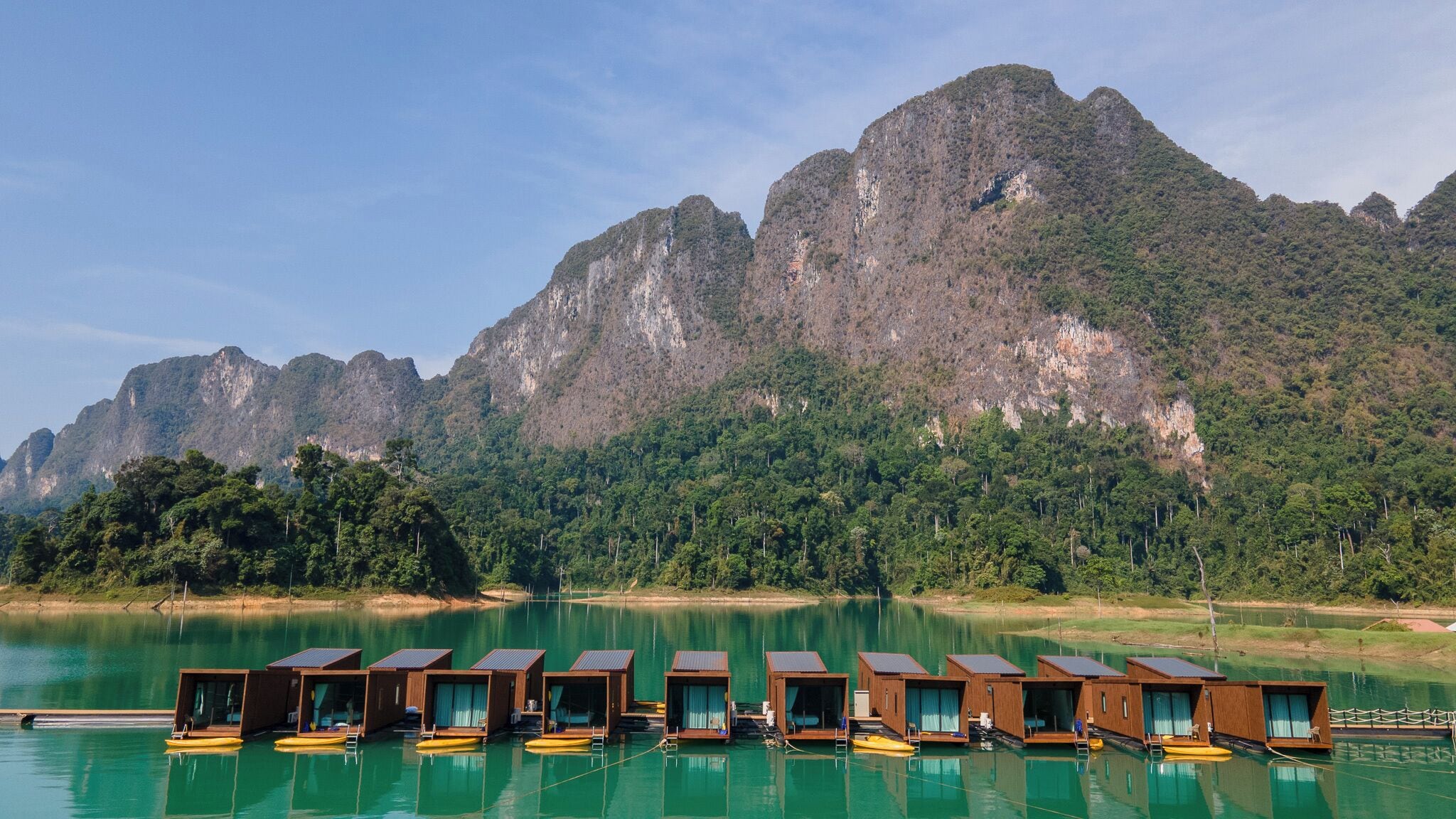 A row of wooden floating bungalows with glass walls, docked in a calm lake, surrounded by lush greenery and mountains.