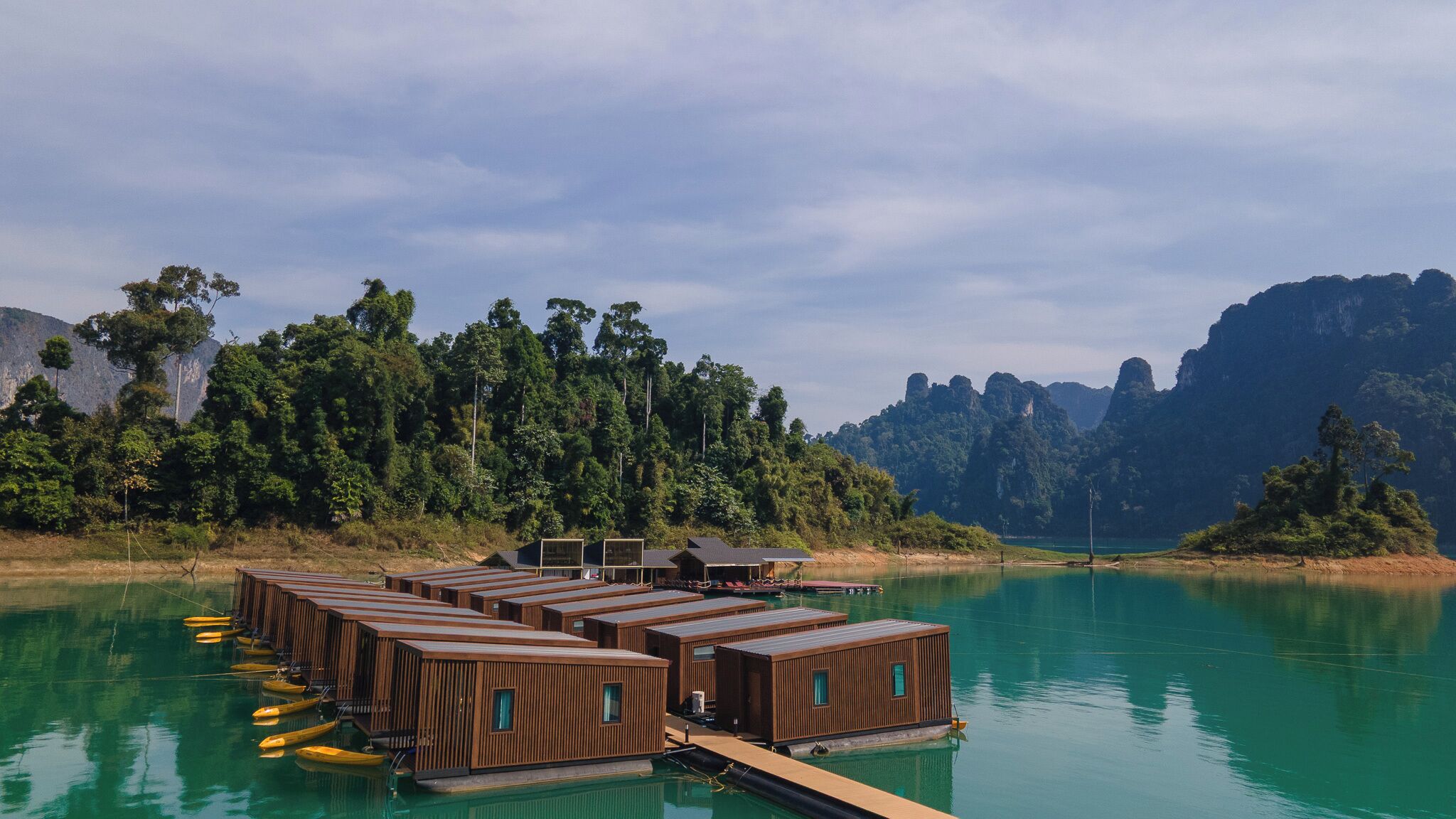 A series of wooden floating structures with windows, docked on a calm body of water, surrounded by dense forest and rocky cliffs.