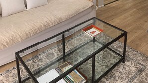 Uma sala de estar com um sofá, uma mesa de centro de vidro, uma luminária e uma cozinha ao fundo.