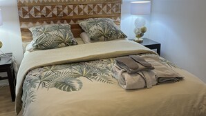 Cama bem arrumada com colcha estampada, travesseiros e toalha dobrada. Há um abajur e um espelho decorativo na parede.