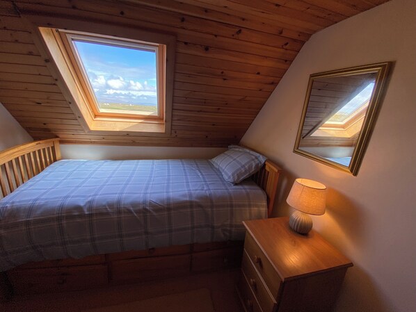 Un dormitorio con techo inclinado, tragaluz, cama con colcha de cuadros, lámpara de noche y cómoda.