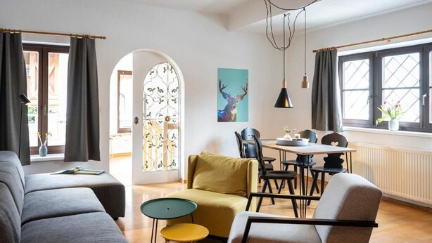 A modern living room with a sofa, a yellow armchair, a dining table with chairs, and a large window with curtains. - Das Nordberg Apartments (Garmisch-Partenkirchen)