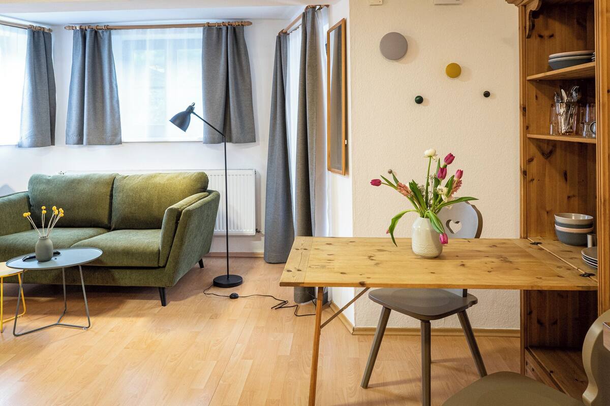 Tunnelmallinen olohuone, jossa on vihreä sohva, puinen ruokapöytä ja maljakko täynnä kukkia.