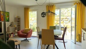 A modern living room with a dining area, a bookshelf, and a large window with yellow curtains.