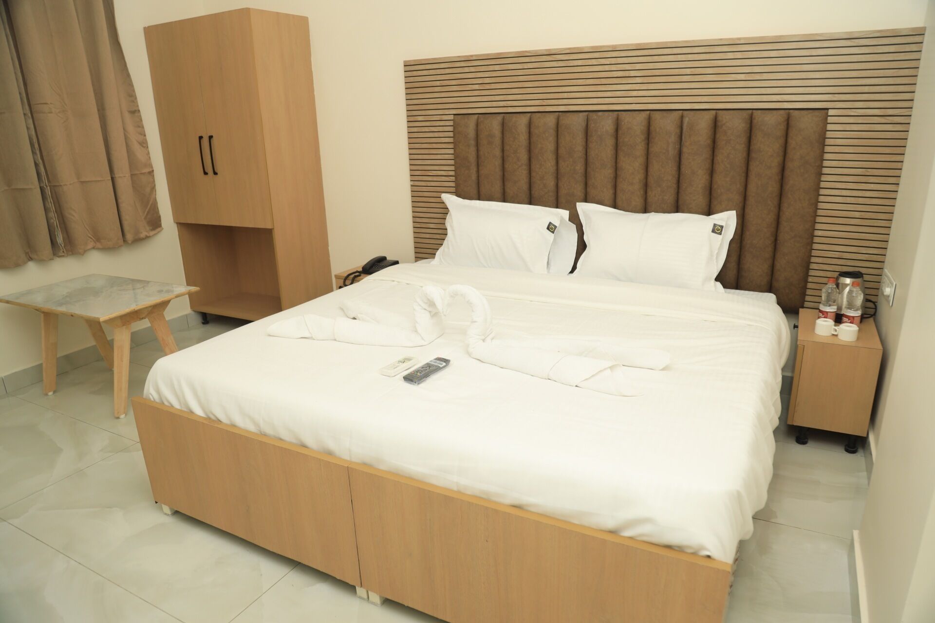 A hotel room with a bed, a wooden wardrobe, a bedside table with a phone, and a small table with a remote control.