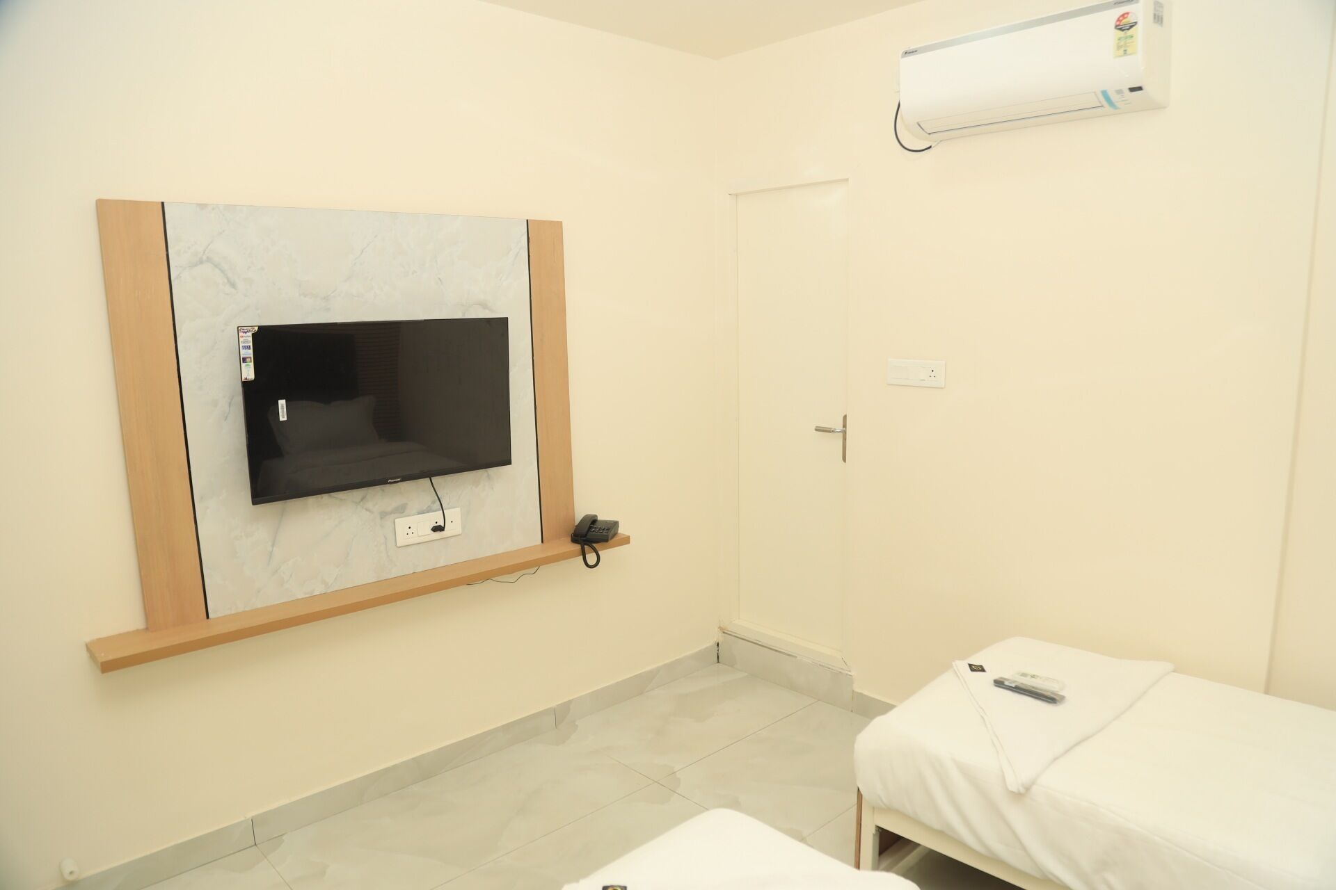 A hotel room with a flat-screen TV mounted on a marble wall, a bedside table with a remote control, and a white bed.