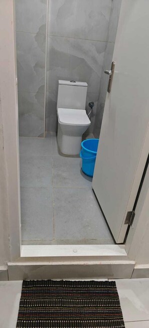 A small, enclosed bathroom with a toilet, a blue bucket, and a drain grate. - Varrahi Exotica  (Hyderabad)