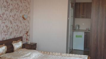 A bedroom with a bed, nightstand, and a small kitchenette area with a refrigerator and a kettle.