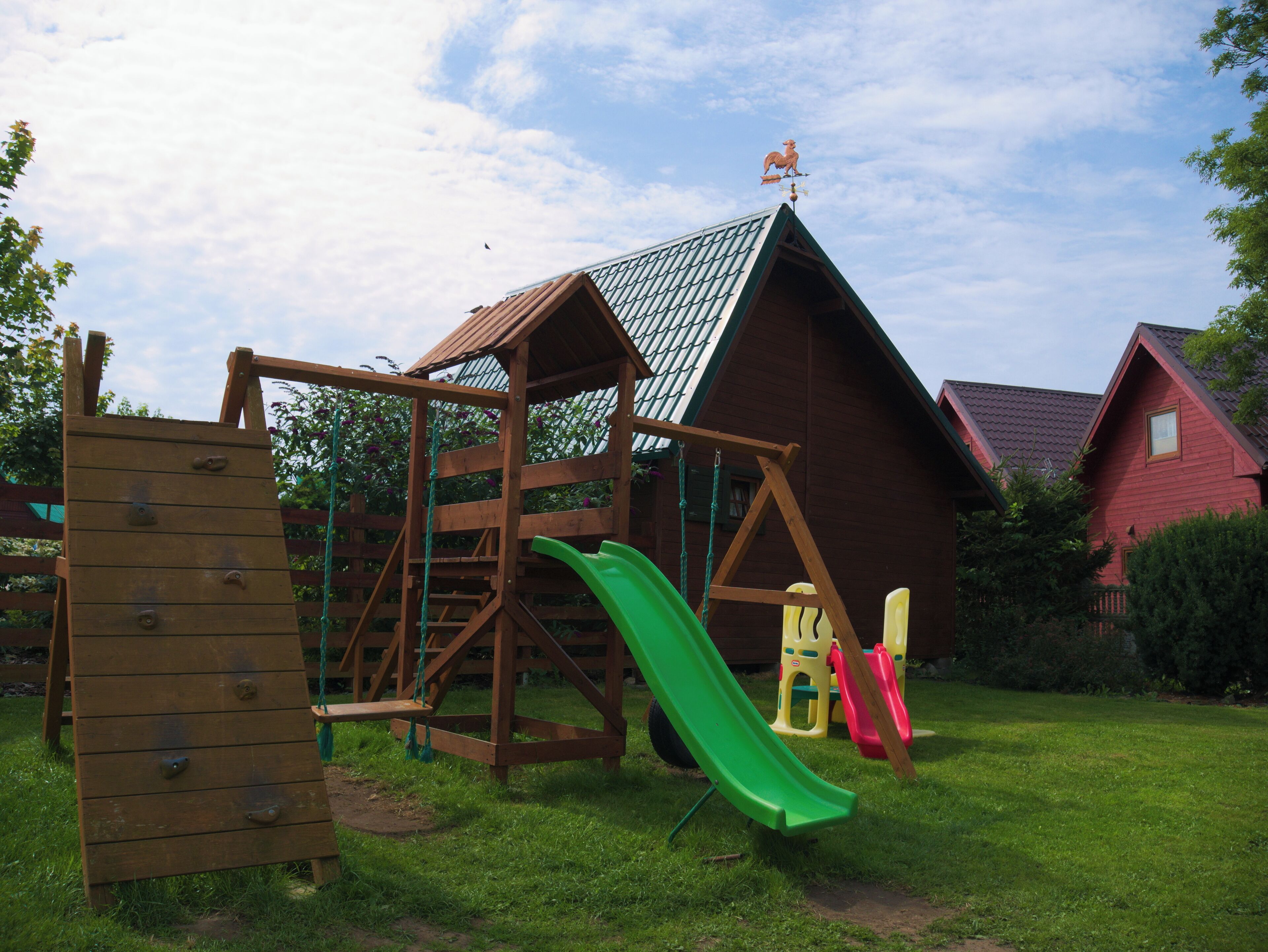 A wooden play structure with a slide and climbing wall, set in a grassy yard with a red-roofed building in the background.