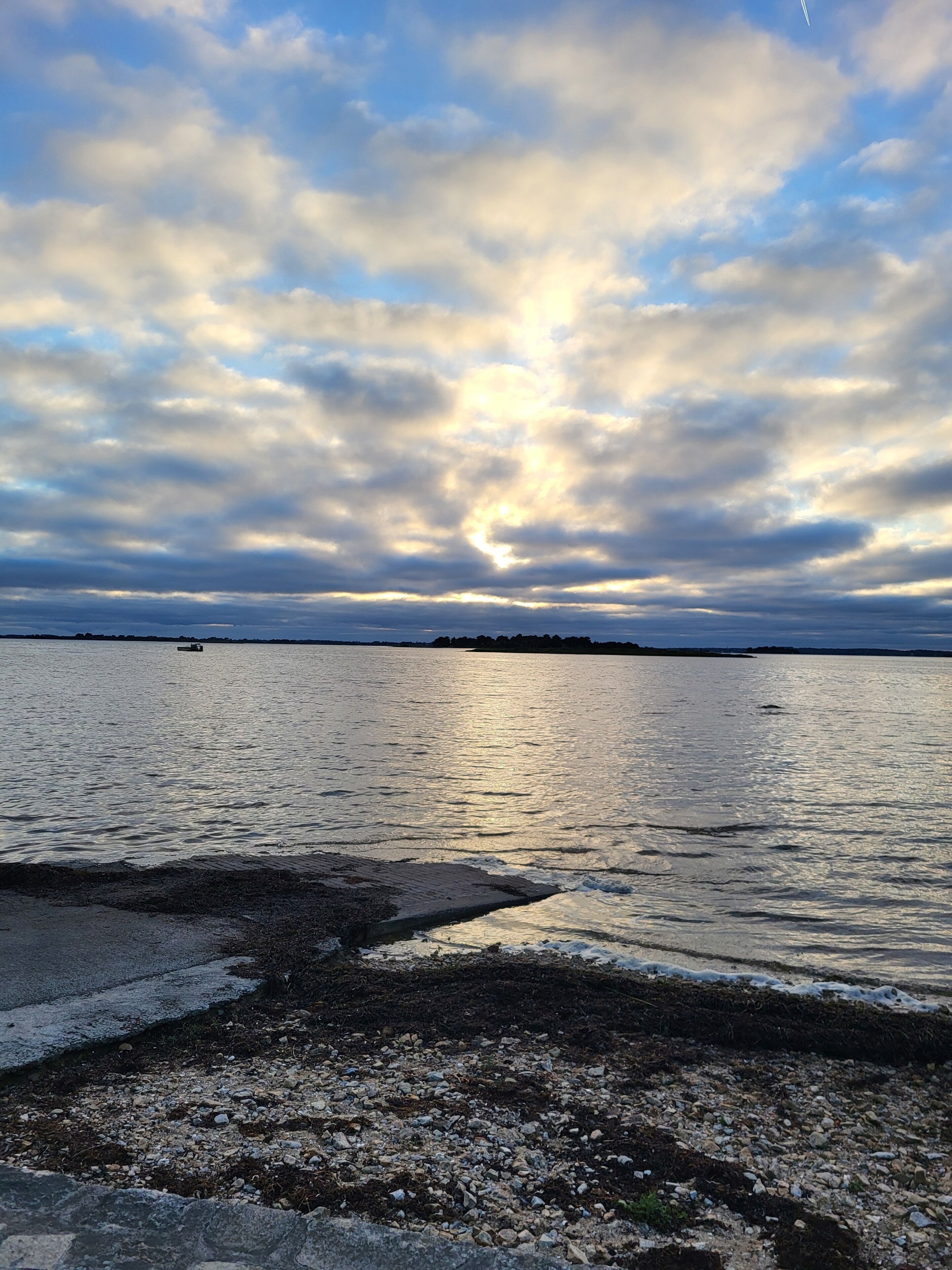 A coastal scene with a rocky shoreline, calm water, and a distant landmass under a cloudy sky.
