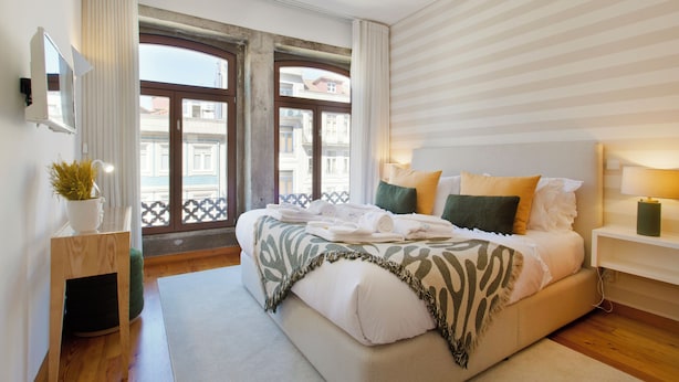A modern bedroom with a large bed, a bedside table with a lamp, a potted plant, and a view of buildings through large windows. - Boutique Rentals-SãO*bEnTo Ribeira Apt (Porto)