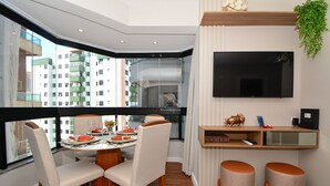 A dining area with a table set for four, a television mounted on the wall, and a balcony with a view of buildings.