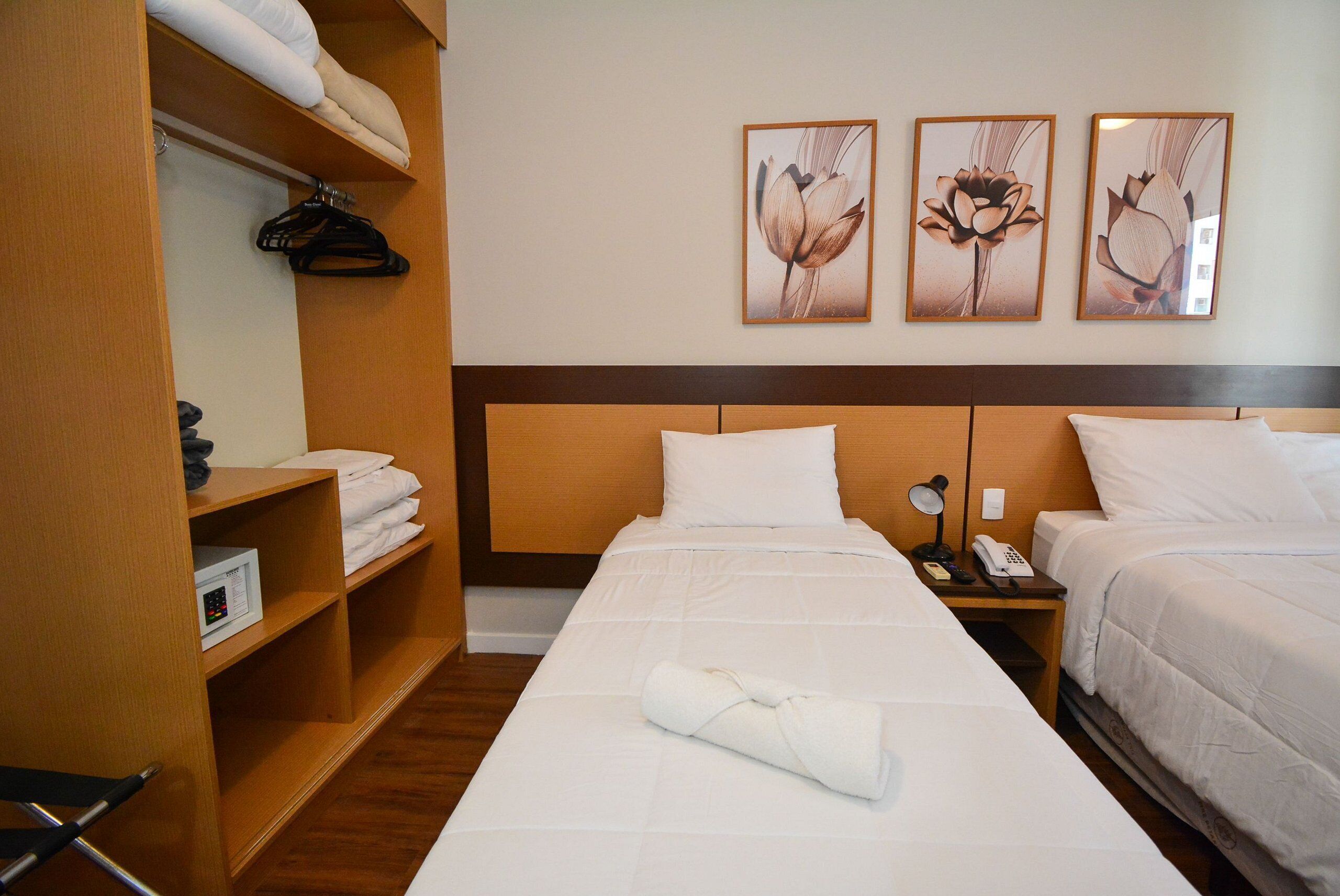 Hotelkamer met een groot bed, nachtkastje met lamp, kledingkast en drie ingelijste bloemstillevens aan de muur.