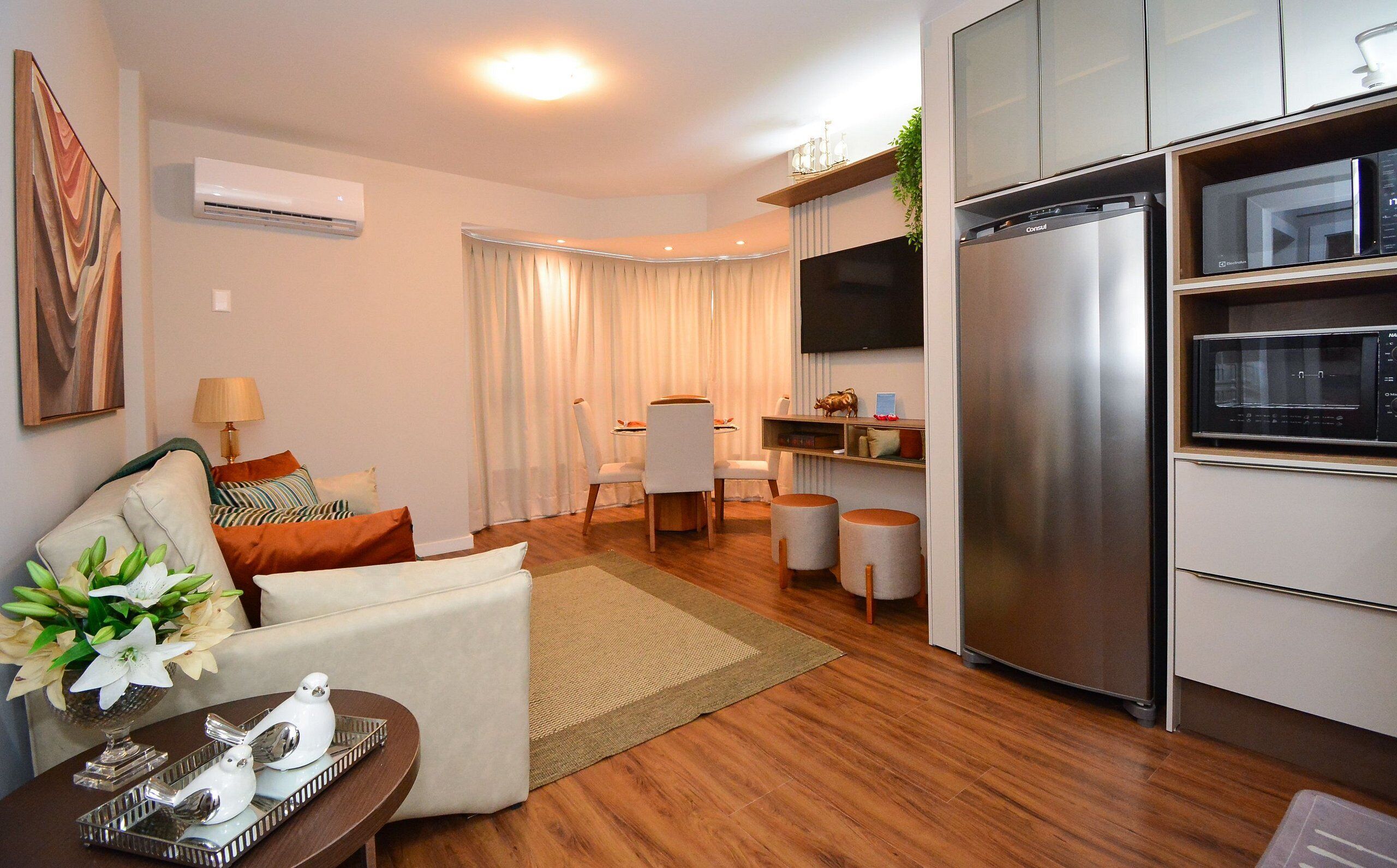 A modern living room with a sofa, a dining area with chairs, a kitchen with a refrigerator and microwave, and a flat-screen TV mounted on the wall.