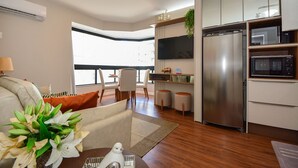 A modern kitchen with a stainless steel refrigerator, microwave, and built-in oven. There is a dining area with a table and chairs, and a living room with a sofa and a decorative plant.
