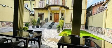 A courtyard with a glass table and chairs, surrounded by buildings with a central staircase and a green lawn.