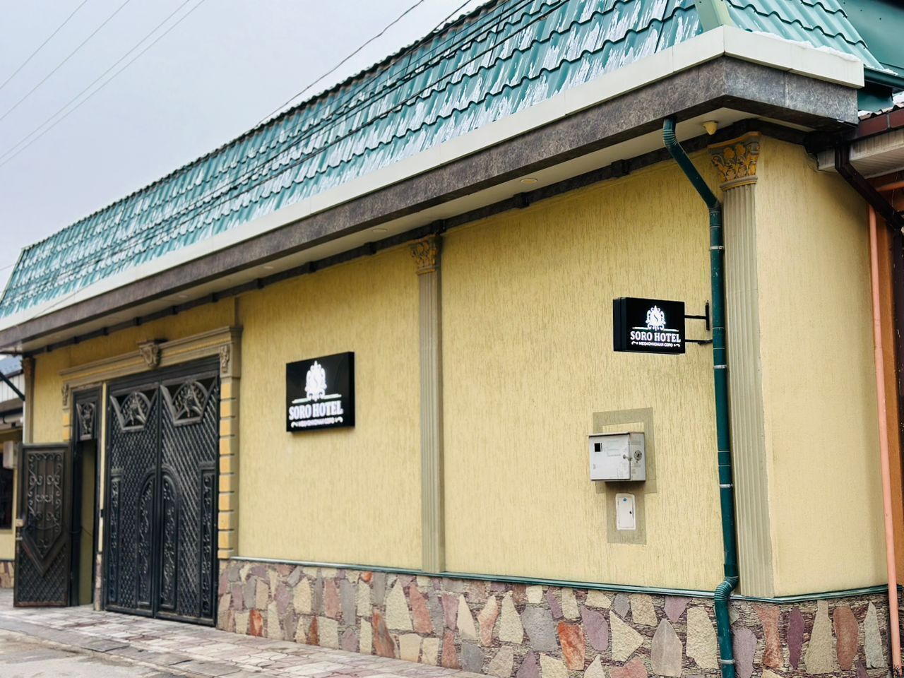 A building with a yellow facade, two black doors, and a green roof. There are signs for 'Soro Hotel' on the wall.