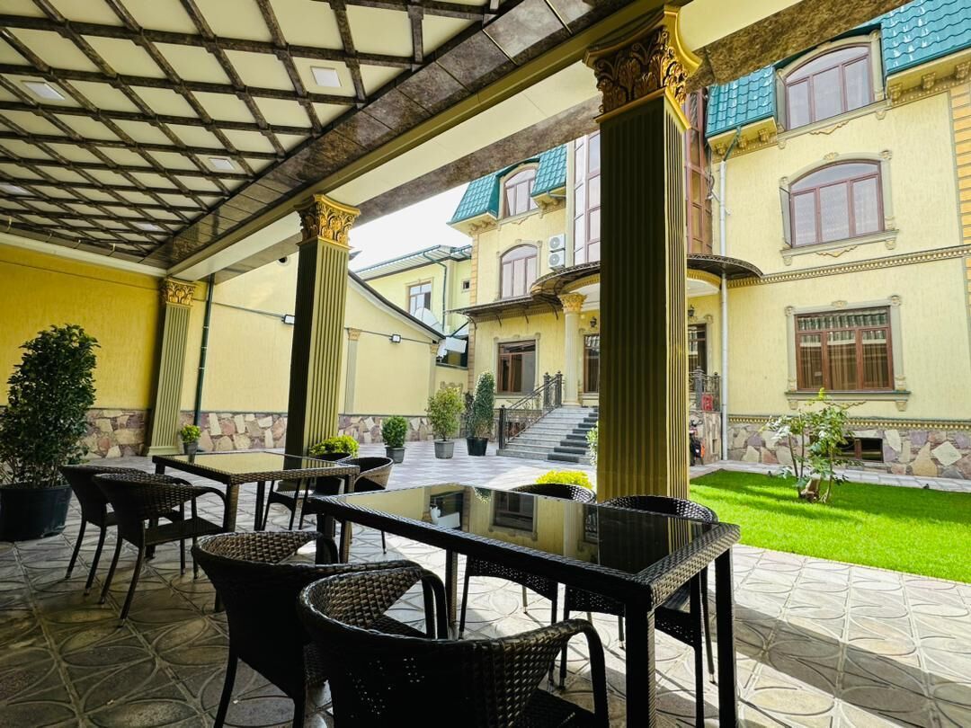 A covered outdoor area with wicker furniture and a glass table, surrounded by a yellow building with a stone facade and a staircase leading to an upper level.