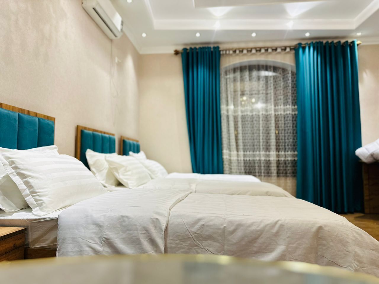 A hotel room with two beds, a wooden headboard, a nightstand, and a window with blue curtains.
