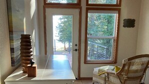 A kitchen with a white countertop, a wooden block, and a wicker chair. There are two windows with views of trees and a deck. - Beautiful Lake Front Home on Lake Almanor. (Westwood)