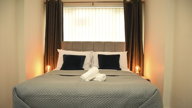 Una cama bien hecha con un edredón gris, dos almohadas y una toalla enrollada sobre el cabecero.