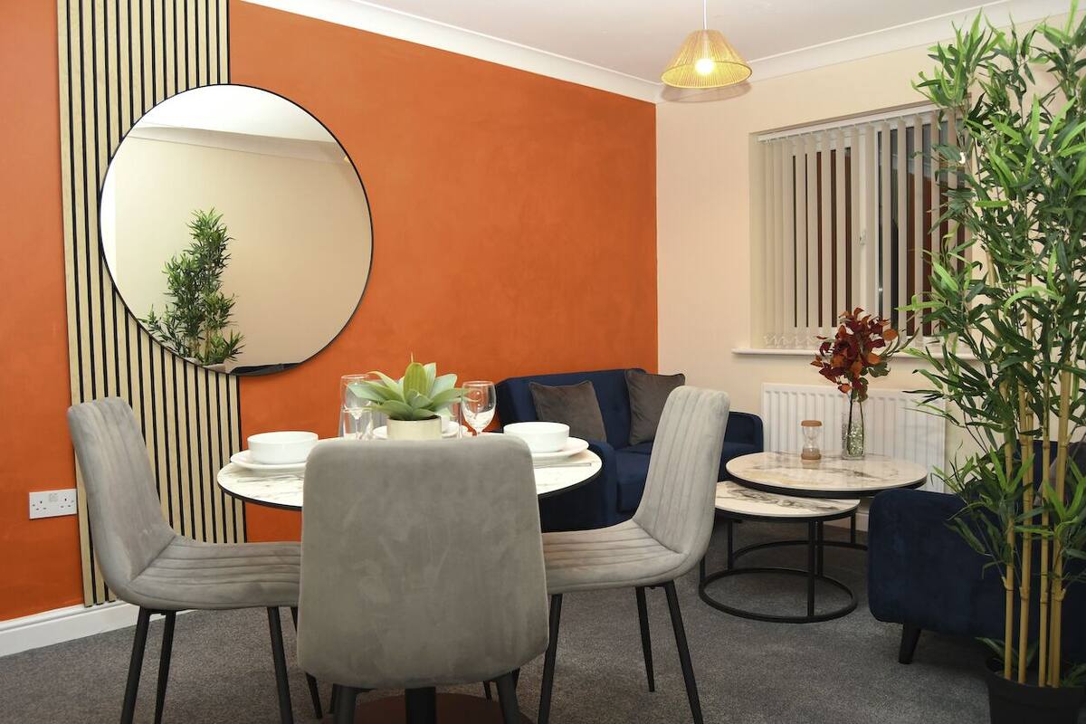 A dining area with a round table, grey upholstered chairs, a blue sofa, a large mirror, and a potted plant.