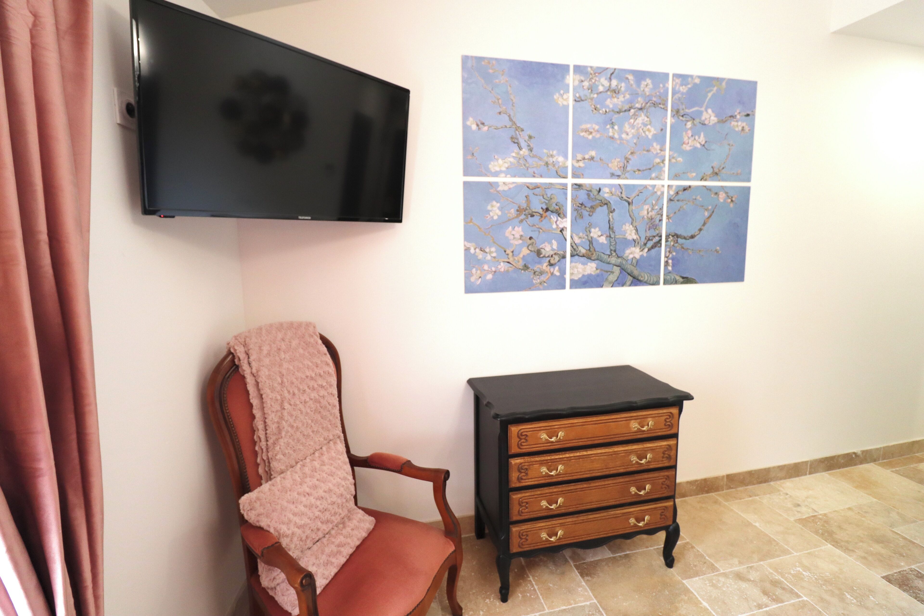A room with a flat-screen TV mounted on the wall, a wooden chair with a blanket, and a dresser with drawers.