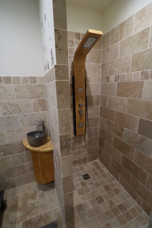 Un área de ducha pequeña con azulejos, un banco de madera y un lavabo tipo cuenco.