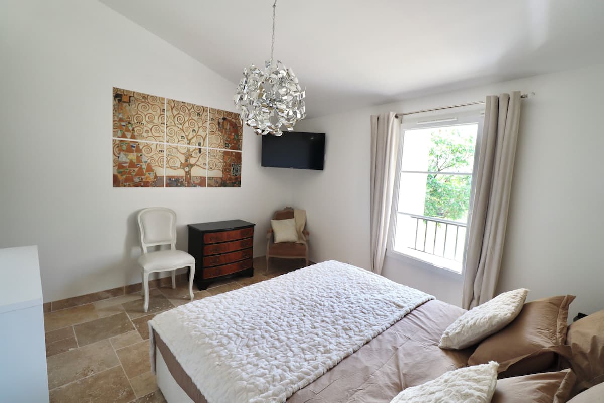 A bedroom with a large bed, a television mounted on the wall, a chair, a dresser, and a window with curtains.