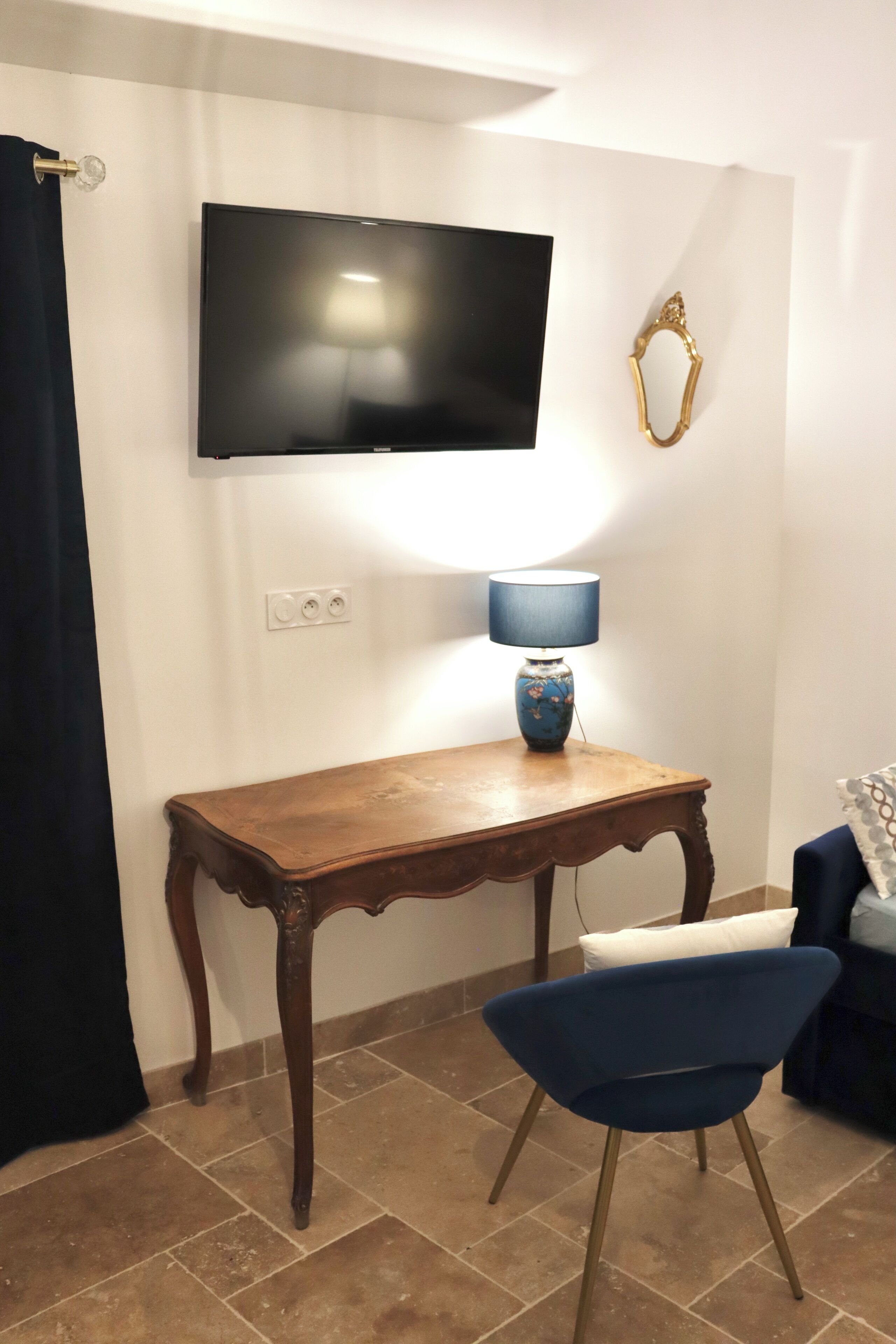 Une pièce avec une table en bois, une chaise bleue, un téléviseur fixé au mur et une lampe sur la table.