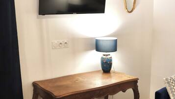 Une pièce avec une table en bois, une chaise bleue, un téléviseur fixé au mur et une lampe sur la table.