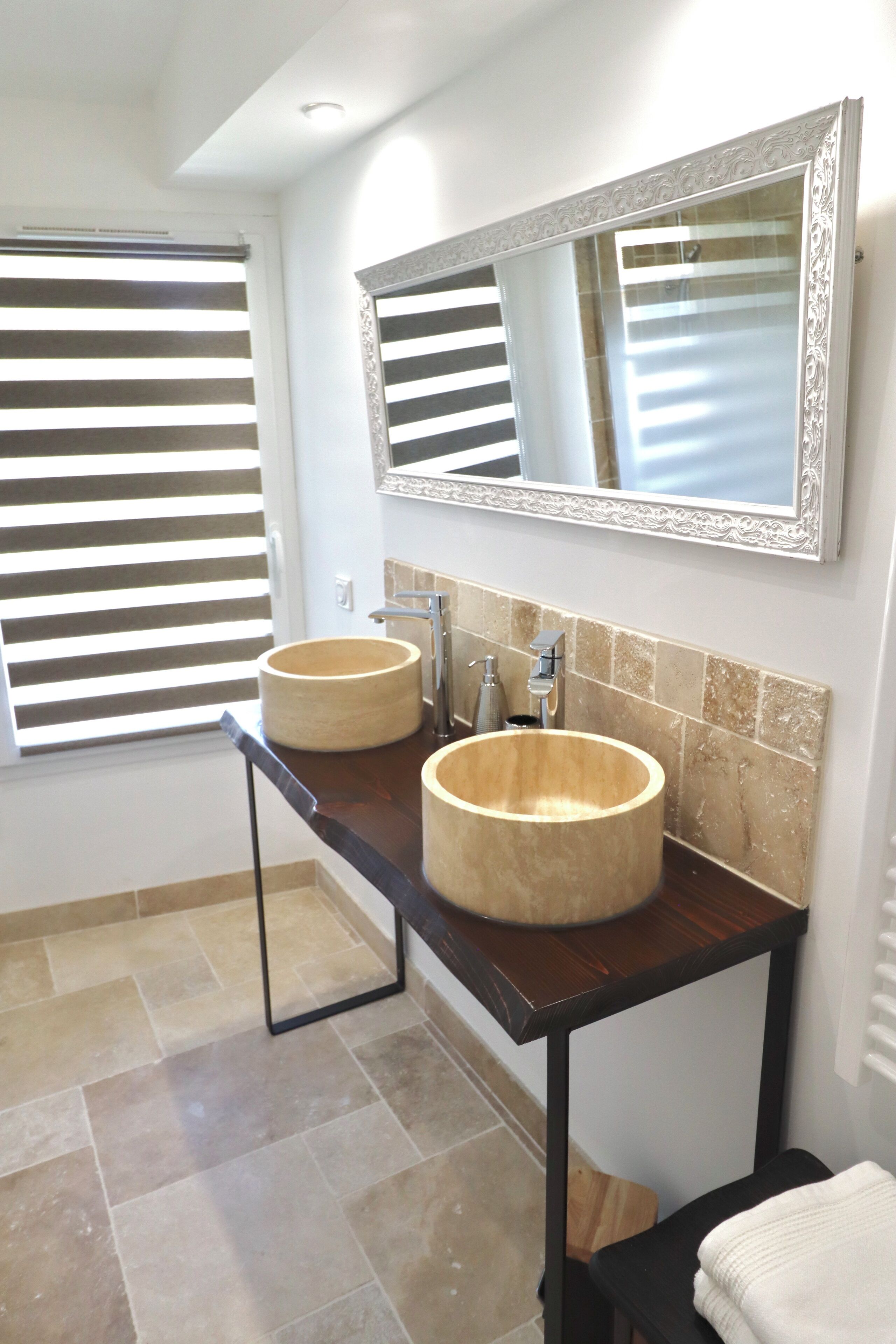 A bathroom with two round sinks, a mirror with an ornate frame, and a window with horizontal blinds.