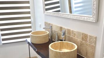 A bathroom with two round sinks, a mirror with an ornate frame, and a window with horizontal blinds.