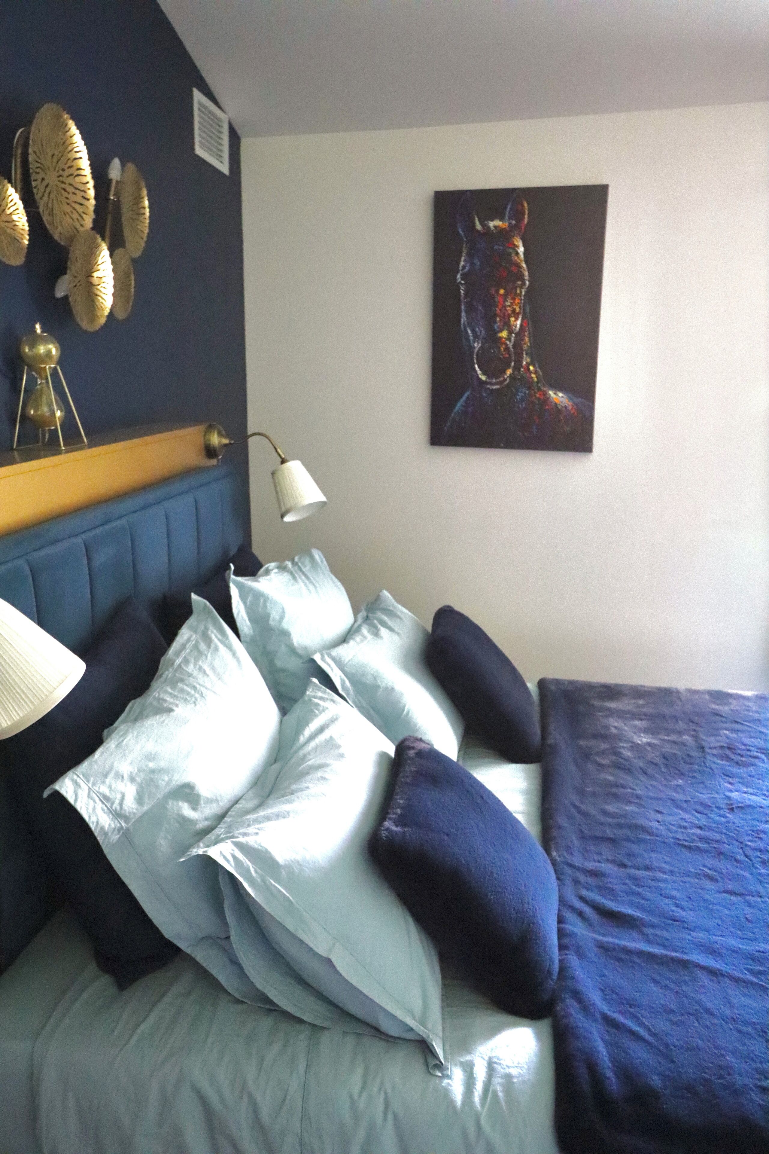 A bedroom with a blue headboard, a bed with light blue bedding, and a painting of a horse on the wall.