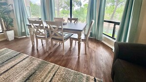 A dining area with a wooden table and chairs, large windows with curtains, and a ceiling fan.