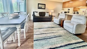 A modern living room with a dining table, sofa, and armchair, a kitchen area with appliances, and a large window.