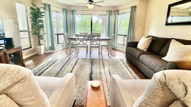A living room with a dining area, a ceiling fan, and large windows.