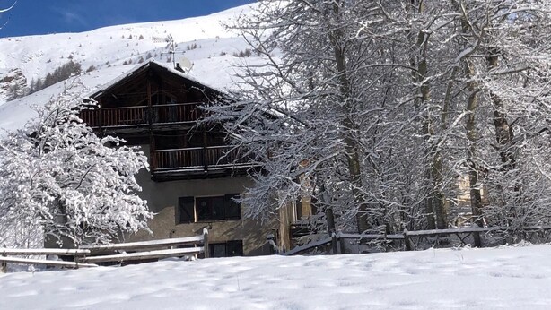 Una xalet de fusta amb un balcó, envoltat d'arbres coberts de neu i un cel blau clar.