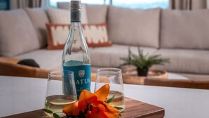 Une bouteille de vin Matua, deux verres et une fleur d’oranger sur un plateau en bois dans un salon donnant sur l’océan.