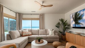 Un salon moderne avec un grand canapé d’angle, une table basse ronde et deux fauteuils. On y trouve un ventilateur de plafond, une grande fenêtre avec des rideaux et un téléviseur fixé au mur.
