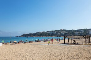 Una playa con gente tomando el sol y caminando, una orilla arenosa y edificios al fondo.