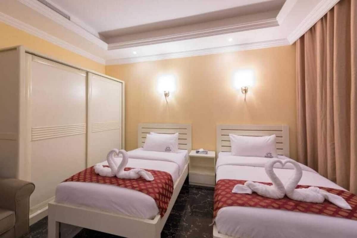 A hotel room with two beds, each with a heart-shaped towel arrangement. There are two bedside tables, a wardrobe, and a chair.