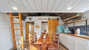 A wooden dining table with chairs, a ladder, and a kitchen area with shelves and appliances.