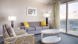 Camera d'albergo con un divano grigio, una poltrona con motivi decorativi e un tavolino rotondo. Si vede una finestra con vista sulla città.