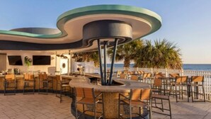Un'area salotto esterna con struttura a tettoia moderna e curvilinea, circondata da sedie e tavoli, con vista sulla spiaggia.