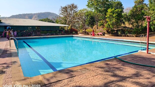 A swimming pool with a tiled deck, surrounded by trees and a building with a green roof. - Green Hills Resort (Vasai)