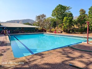 A swimming pool with a tiled deck, surrounded by trees and a building with a green roof. - Green Hills Resort (Vasai)